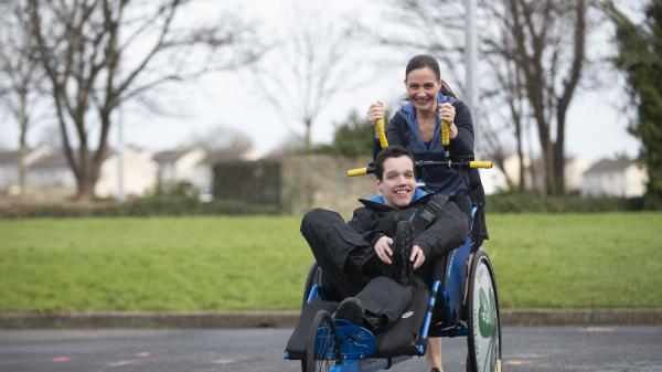 Female runs through the park pushing an adapted wheelchair in front of her with her son